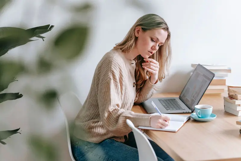 Vrouw maakt aantekeningen in een notitieboek naast haar laptop en een kopje koffie.