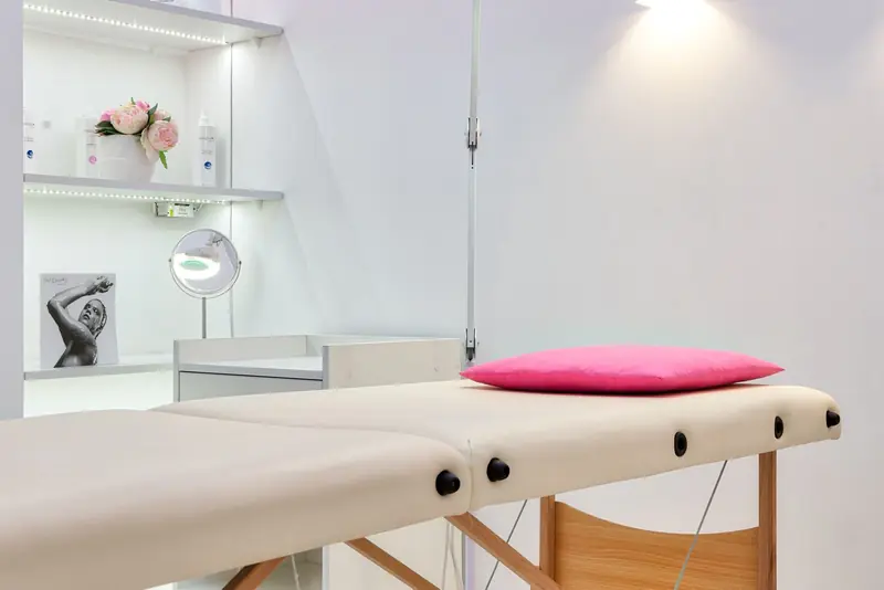Lichte behandelkamer met een behandelbank, een roze kussen en een loeplamp aan de wand.