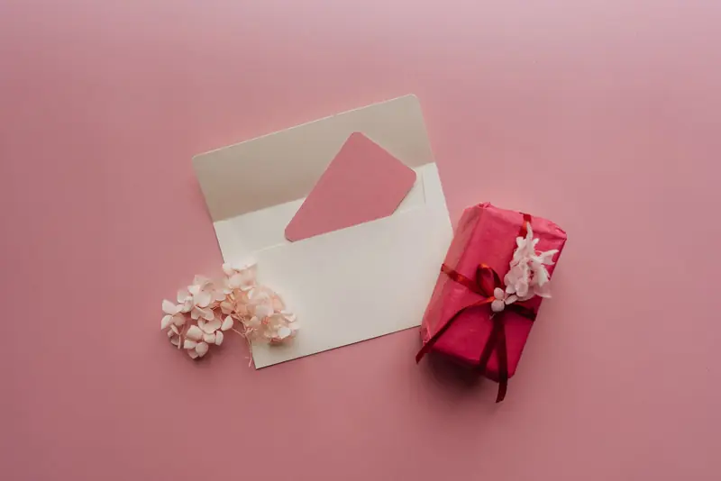 Witte envelop met een roze kaart naast een ingepakt cadeautje op een roze ondergrond.