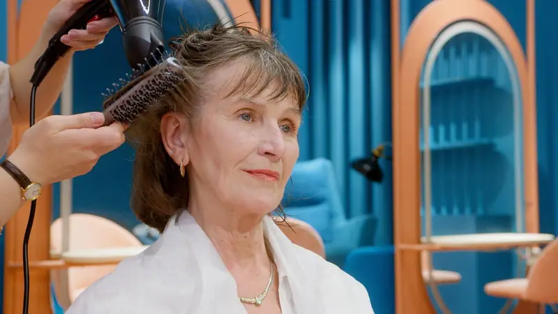 Kapper föhnt het haar van een oudere klant met een ronde borstel in een kleurrijke salon.