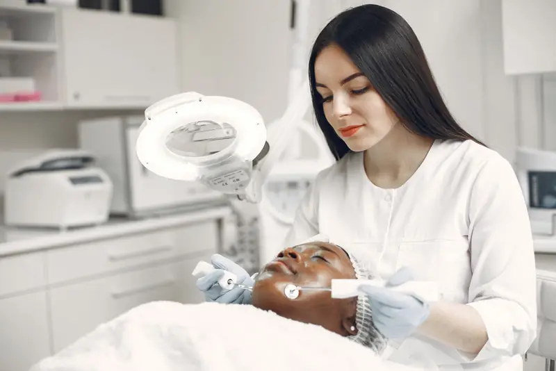 Schoonheidsspecialiste geeft een huidbehandeling onder een loeplamp aan een klant met haarnetje.