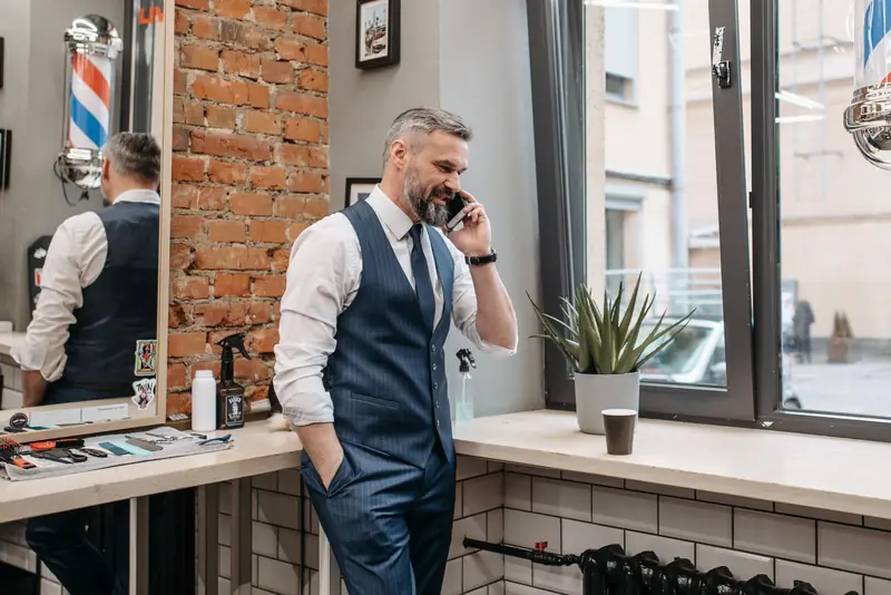 Kapper in gilet belt met zijn telefoon bij het raam van een lichte barbershop.