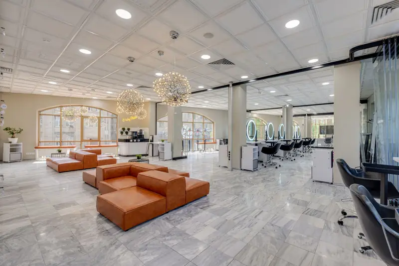 Lichte kapsalon met marmeren vloer, cognackleurige loungebank en een rij kappersstoelen voor ronde spiegels.
