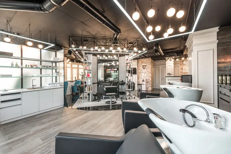 Moderne kapsalon met wasbakken, zwarte stoelen en een plafond vol lampen.