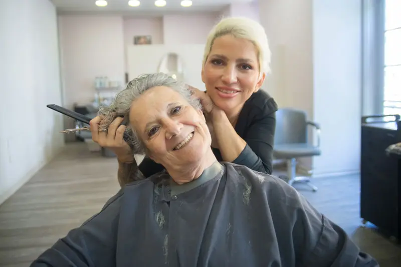 Kapster en oudere klant lachen samen in de spiegel tijdens een knipbeurt in een lichte salon.