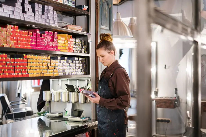 Kapster kijkt op haar telefoon naast een wand vol haarverf in een drukke kapsalon.