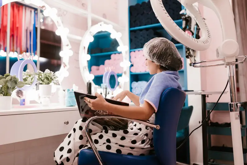 Klant met haarnetje bekijkt een tablet in een salon met verlichte spiegels.