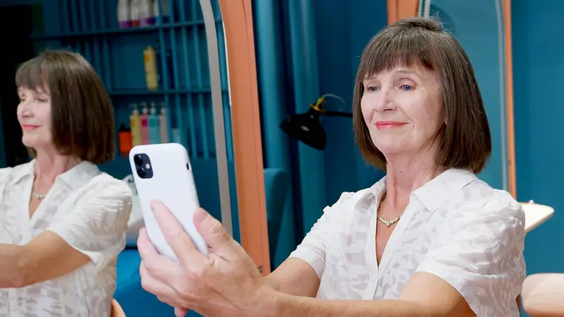 Oudere vrouw maakt een selfie met haar telefoon in een salon met blauwe kasten.