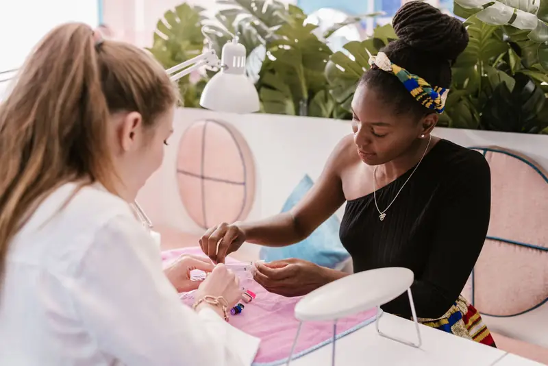 Nagelstyliste werkt aan de nagels van een klant aan een nageltafel met planten erachter.