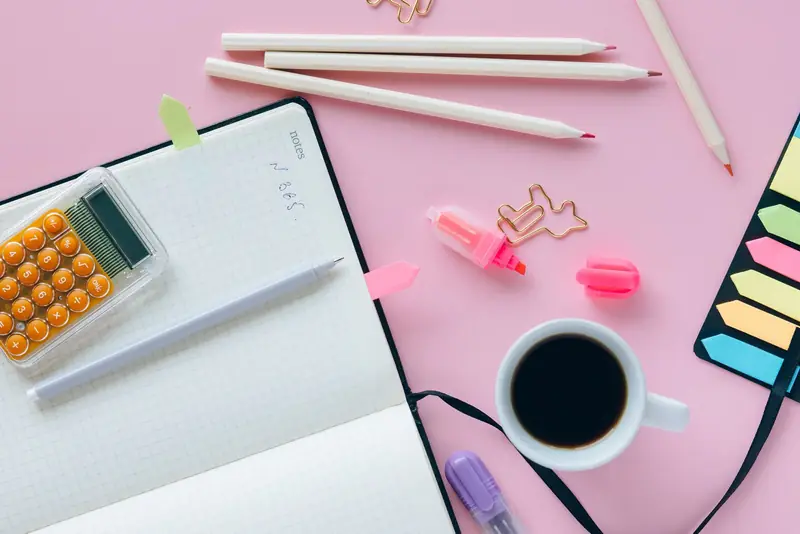 Notitieboek, rekenmachine, potloden en een kop koffie op een roze bureau.