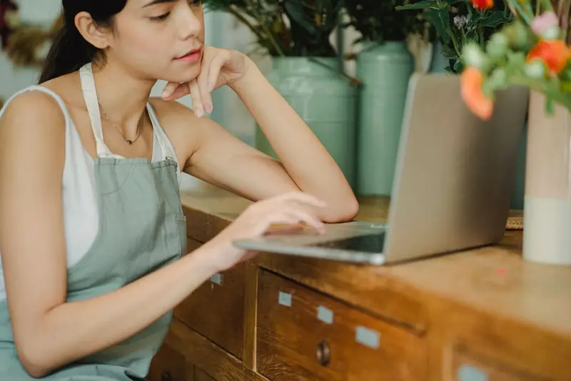 Ondernemer in schort leunt op een houten werkbank en werkt op haar laptop tussen bloemen.