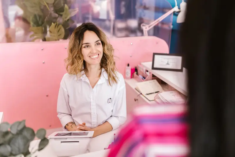 Lachende receptioniste achter een roze balie met kassa ontvangt een klant in een salon.