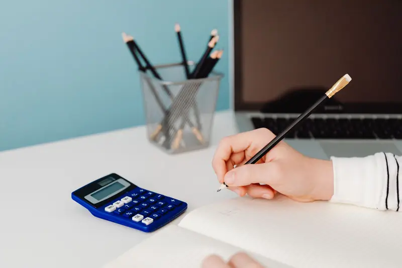 Hand schrijft met potlood in een notitieboek naast een blauwe rekenmachine op een wit bureau.