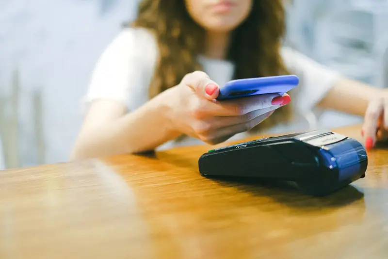 Klant betaalt contactloos met haar telefoon bij een betaalterminal op een houten balie.