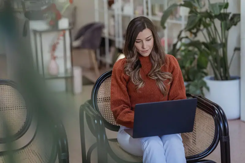 Vrouw in rode trui werkt op haar laptop in een rieten stoel tussen planten.