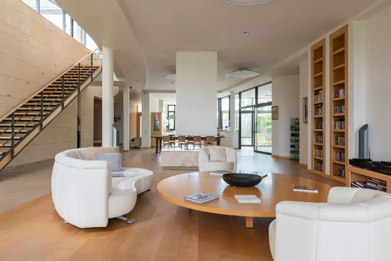Ruime lichte wachtruimte met witte fauteuils, een ronde houten tafel en een trap naar boven.